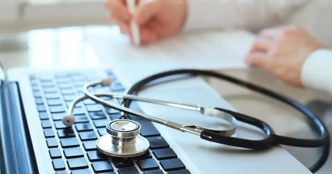 : image of a stethoscope sitting on a laptop while a physician does paperwork in the background