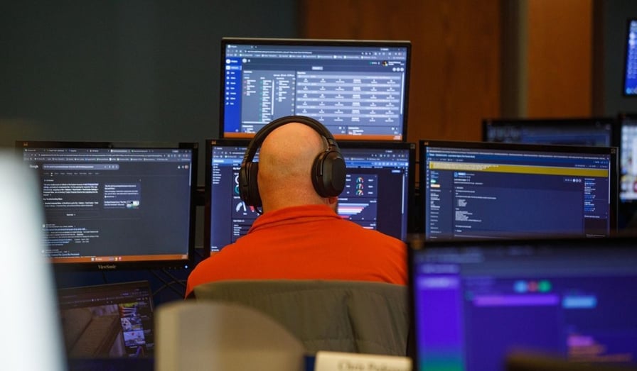 An IT security professional wearing headphones monitors real-time network data and cybersecurity dashboards across multiple computer screens in a security operations center.