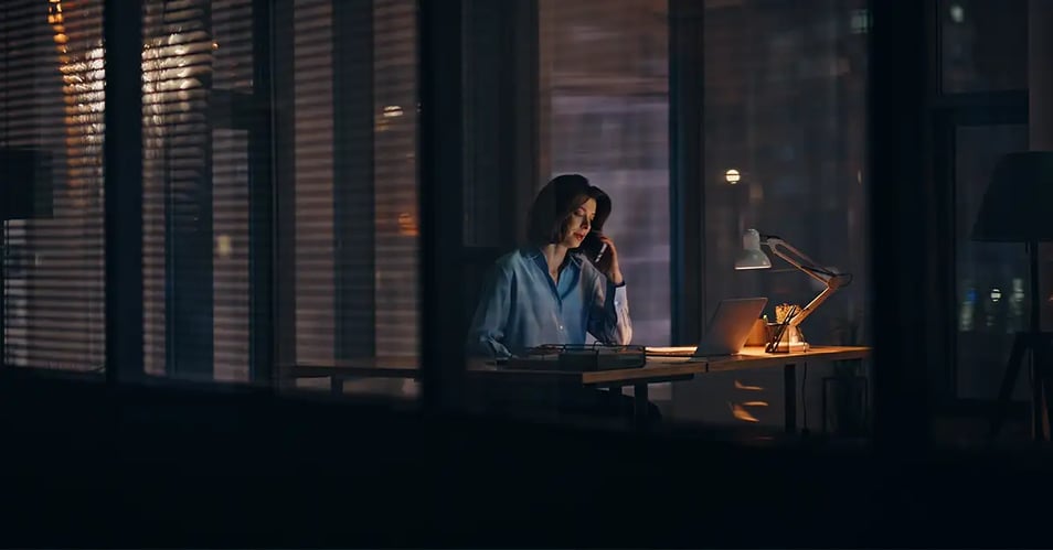 Woman working alone in a bank office after hours while speaking on the phone during an IT emergency.