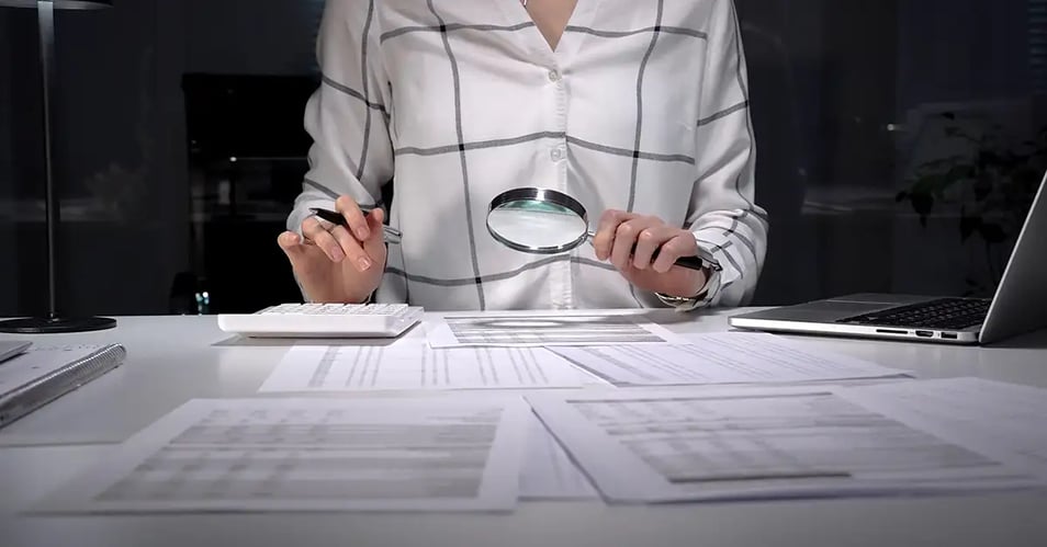 A professional using a magnifying glass to review audit documents on a desk during bank audit preparation.