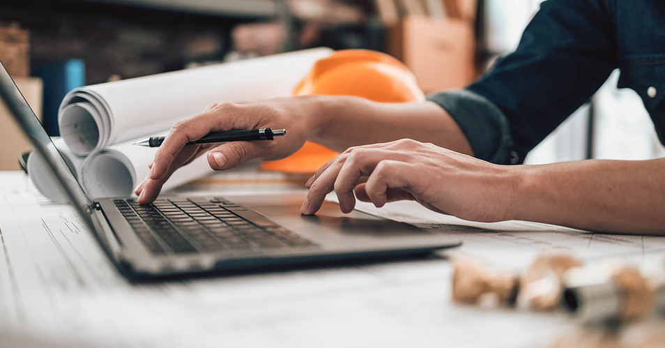 A construction professional works on a laptop at a desk with rolled blueprints, an orange hard hat, and a pen in hand, illustrating technology use on a construction project.