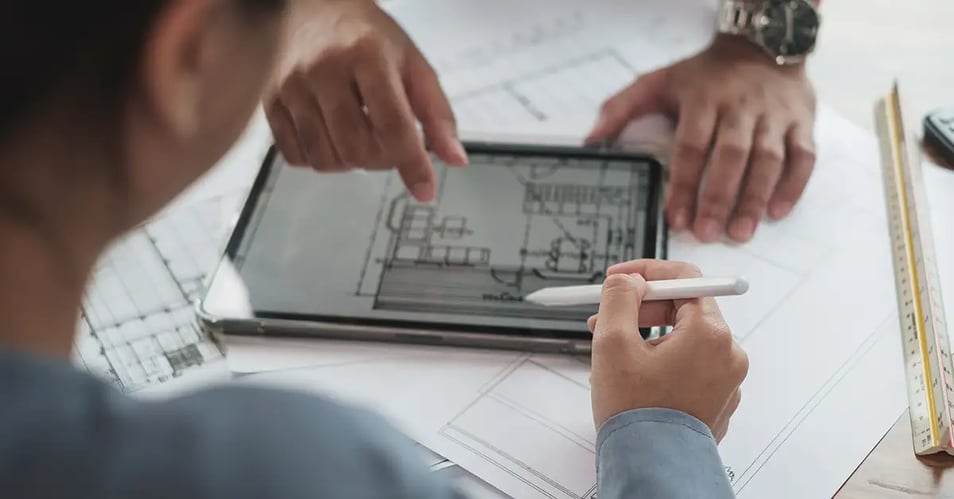 Two people reviewing a construction floor plan on a tablet surrounded by printed blueprints, illustrating IT support for distributed construction teams.