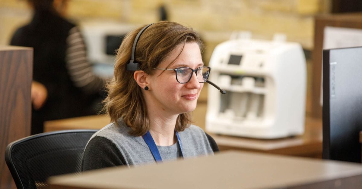 an image of a local community bank teller sitting at the counter looking at her computer with her headset on with a money counter blurred in the background