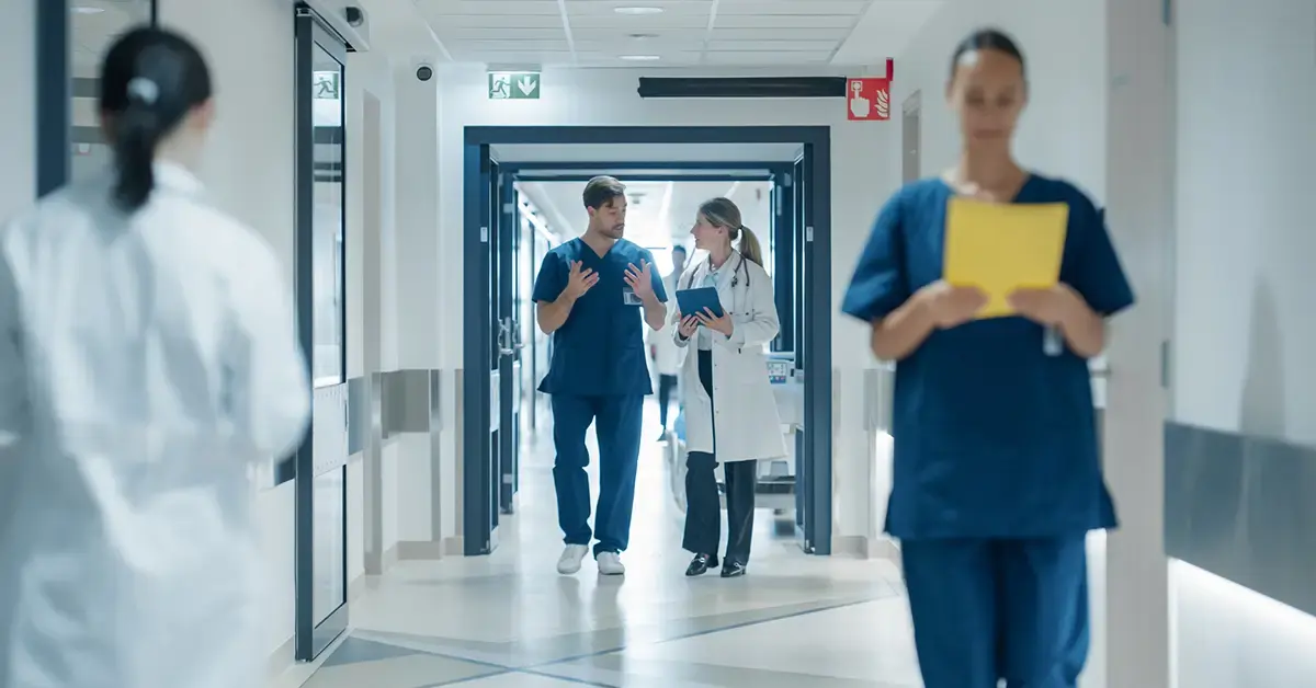 Hospital corridor with medical staff walking; a doctor and nurse discuss patient information at center, representing medical device cybersecurity in a modern healthcare setting.